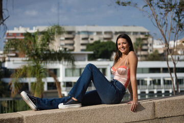 Young beautiful woman with straight brown hair, orange top and jeans, sitting on a stone wall, looking at the camera, sunbathing relaxed and happy. Concept fashion, beauty, trend, relax, millennial.