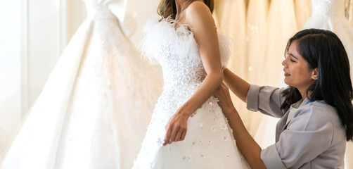 Portrait of smiling happy asian fashion bride woman talk and try on wedding dress with woman shop owner for pre wedding at wedding interior fashion studio.