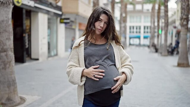 Young pregnant woman standing with serious expression touching belly at street