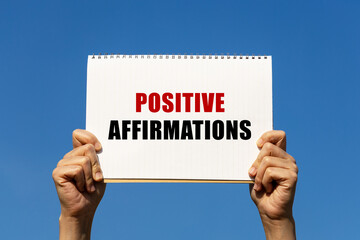 Positive affirmations text on notebook paper held by 2 hands with isolated blue sky background....