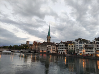Fototapeta na wymiar Zurich, Switzerland. River Limmat. Sunset sky, dark clouds, evening time. Orange lights, sundown. Old city centre, clock tower, urban landscape.