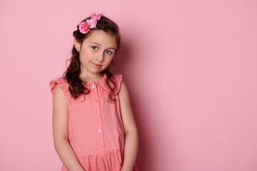 Obraz na płótnie Canvas Adorable beautiful Caucasian noble little child girl wearing stylish pink dress and hairstyle with roses, smiles looking at camera with insightful gaze, isolated pink backdrop. Copy advertising space