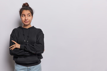 Studio shot of lovely dark haired woman keeps arms folded concentrated aside thoughtfully tries to find solution in mind dressed in casual black sweatshirt and jeans isolated over white background.