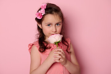 Close-up beauty portrait of a beautiful Caucasian blue eyed little kid girl in pink dress and flowers in hairstyle, sniffing a pink rose flower, smiling, looking at camera, isolated on pink background