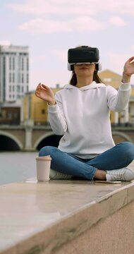 Young woman wearing VR glasses, immersed in a virtual world, while gesturing in front of bustling city skyline. Potential of cutting-edge technology in the heart of the metropolis. Vertical 4k video.