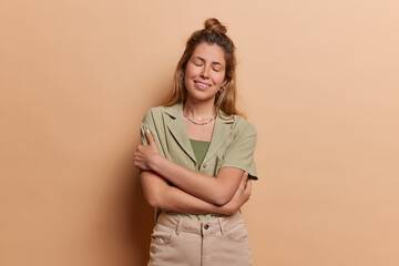 Obraz premium European girl wears shirt trousers and embraces herself with warm smile sstands isolated against beige background emanating sense of inner peace and self acceptance isolated over brown background