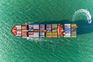 Top view cargo container ship with tug boat carrying container and running on emerald green waters.
