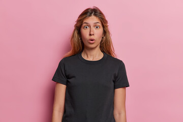 Young European woman expresses utter shock has eyes wide and jaw dropped in disbelief unexpected turn of events has left her completely stunned wears casual black t shirt isolated over pink background