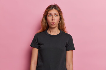 Fototapeta premium Young European woman expresses utter shock has eyes wide and jaw dropped in disbelief unexpected turn of events has left her completely stunned wears casual black t shirt isolated over pink background
