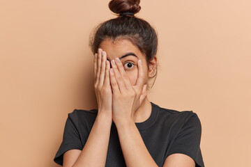Scared Latin woman with hair bun peeks through her fingers fearfully hiding her face unable to bear sight of something terrible witnessing something truly terrible dressed in casual t shirt isolated