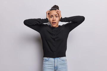 Horizontal shot of Latin girl with dark hair gazes in astonishment her hands on head mesmerized by captivating sight wears black jumper and jeans isolated over white background. Omg what to do