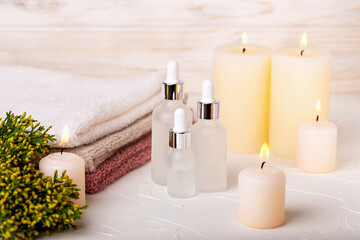 Fototapeta na wymiar White glass dropper bottles with gel or lotion, oil or toner among burning candles. Towels and plant complete concept of spa treatments. Care and rest, beauty and health. Selective focus