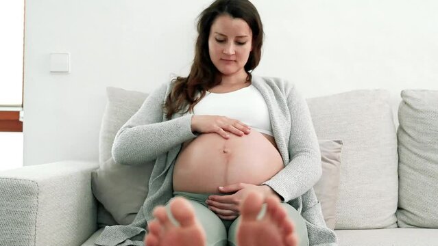 Happy pregnant woman sitting on sofa gently caressing her belly in expectation of baby in last stage of pregnancy. Pregnancy third trimester - week 34. Front view. White background.