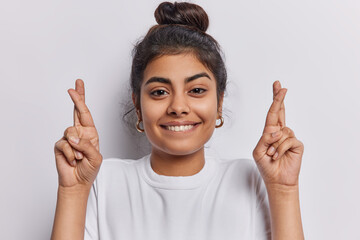 Lovely young Indian woman stands with crossed fingers and hopeful smile radiating strong belief in positive outcomes embodies desire for good fortune capturing essence of optimism wears casual t shirt - obrazy, fototapety, plakaty