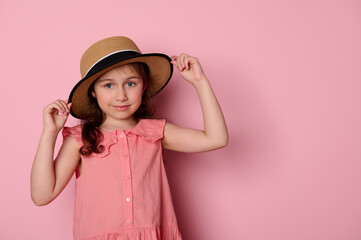 Beautiful Caucasian little child girl 5-6 years old, wearing a stylish pink dress and straw hat, smiling cutely looking at camera, isolated on pink background with copy advertising space. Summer. Kids