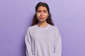 Fototapeta premium Studio shot of pretty dark haired woman purses lips has dissatisfied expression dressed in casual pullover feels frustrated isolated over purple background. Negative human face expressions concept