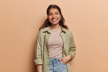 Portrait of cheerful lovely woman with dark hair smiles pleasantly keeps hand in pocket wears shirt and jeans looks directly at camera isolated on brown background. People positive emotions and style