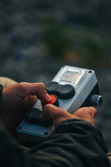 The photo depicts hands holding and pressing the buttons of a control panel. The control panel has several large buttons, including a red button that one of the hands is pressing. The background is bl