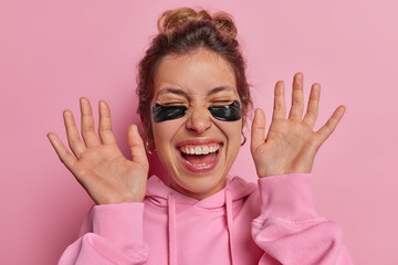 Overjoyed young woman cherishes her skincare routine gracefully applying hydrogel patches keeps palms raised up exclaims loudly wears casual sweatshirt isolated over pink background. Beauty concept