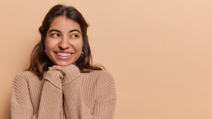 Horizontal shot of pretty dark haired woman looks aside has toothy smile keeps hands under chin dressed in loose knitted jumper poses against brown background copy space for your promotional content © WHstudio Leushin N
