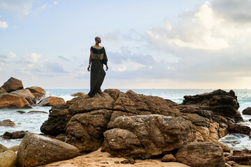 Androgynous black divine person poses on scenic ocean beach. Non-binary ethnic fashion model in...