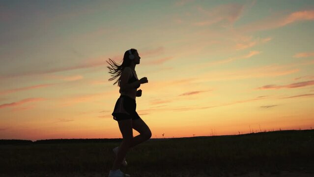 Active young woman runs in summer in park at sunset, listens to music on headphones. Silhouette, beautiful girl in musical headphones, is engaged in fitness, runs in evening. Training run. Teen runner