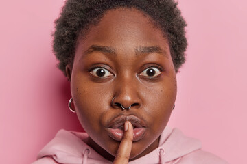 Close up shot of surprised dark skinned young African woman presses finger to lips tells something...