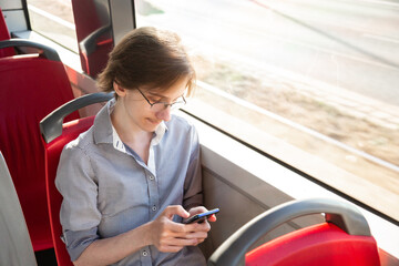 Tennager boy in a tram holding mobile phone in his hand. Guy siting near a window, looks with interest at screen of smartphone, playing mobile games online on smartphone connected to public wifi