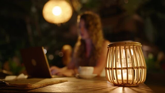 Female freelancer working at night in atmospheric outdoor tropical location. Woman sitting at table outside in dark cafe with warm lights and straw and wooden lamps using laptop. Working late
