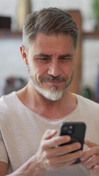 Portrait of happy, confident older man at home using phone, smiling. Mature age, middle age, mid adult casual guy in 50s, bearded, gray hair.