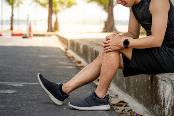 Athlete man feeling knee pain or rheumatoid arthritis while sitting on footpath in the park. Asian...