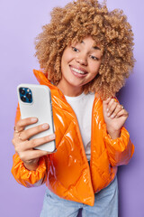 Pretty curly haired woman holds mobile phone makes selfie or online call dressed in orange jacket...