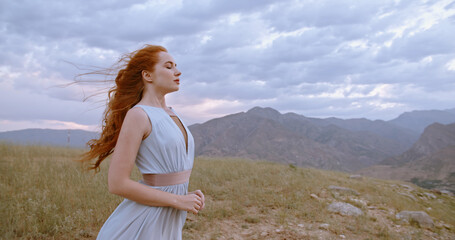 Copy Space. Gorgeous caucasian girl with red hair looking at scenic view from top of a mountain. Wind cinematic blowing hair and white dress - tranquility, peace, freedom.