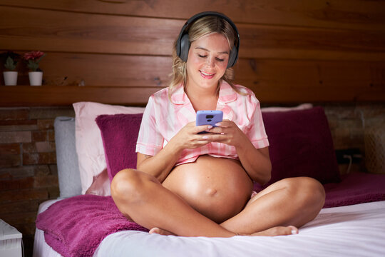 Smiling pregnant woman sitting on bed at home using cell phone. Cheerful mom connected online and listening to music on headphones . Concept of happy people and motherhood.