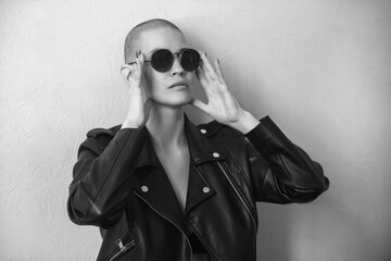 Portrait of a beautiful young woman with a shaved bald head in dark sunglasses and a black leather jacket on a light background. Black and white photo