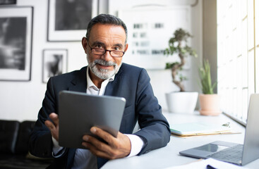 Fototapeta premium Glad confident european senior businessman in suit, glasses uses tablet at table in office