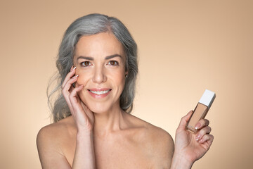 Portrait of beautiful senior european woman with clean glowing skin, holding foundation makeup bottle, beige background