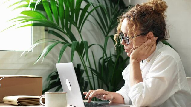 Modern woman entrepreneur doing boring work at laptop. Online job and boredom. People work in indoor home office workplace no happy. Tired and stressed lady using notebook on the table with plant back