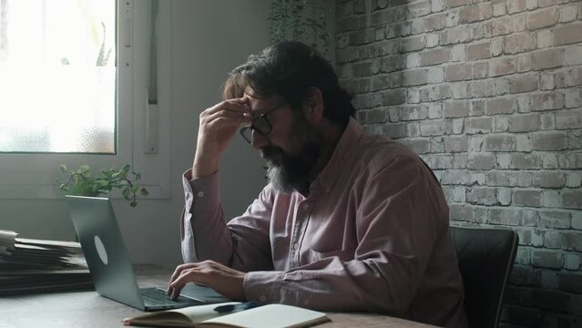 One man alone at the desk using laptop with stress expression and posture. Dark light. Problems and difficulty. People painful for migraine headache using computer indoor. Unhappy worker with notebook