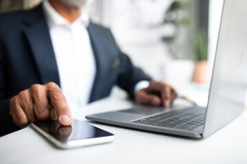 Fototapeta premium European senior businessman in suit sits at table with computer, typing on smartphone with blank screen