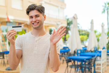 Young handsome man holding a credit card at outdoors saluting with hand with happy expression