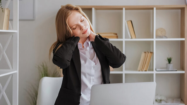 Neck exercise. Sitting work. Office syndrome. Tensed young woman in business wear before computer massaging spine with closed eyes in light room.