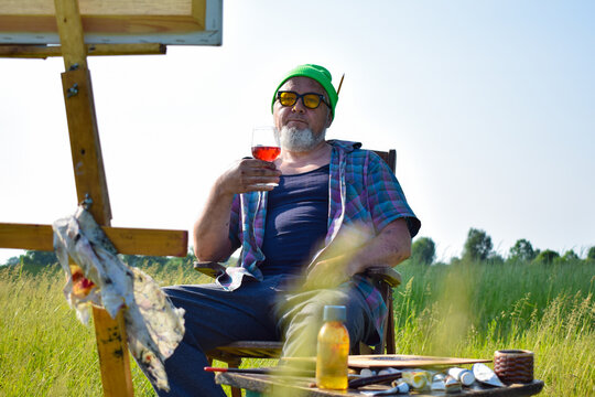 Offbeat mature artist relaxing after completion artwork at field, drinks wine looking at picture sitting on chair outdoors in morning sunshine. Canvas on wooden easel painting tools materials supplies