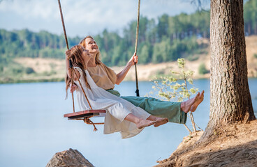 Mom swings with her daughter on a rope swing against the backdrop of a forest lake reflecting the sky. Family concept