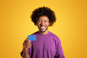 Cheerful black adult curly man in purple t-shirt shows credit card, isolated on orange background, studio