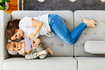 Woman and her little son lying on cozy sofa with smartphone, above view