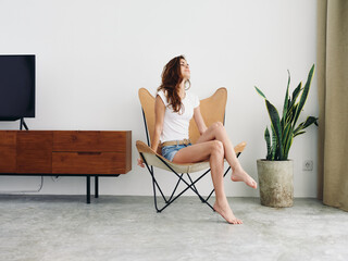 Fototapeta na wymiar Woman sitting in leather armchair smile with teeth Lifestyle in white T-shirt and blue denim shorts, resting at home stylish modern interior with white walls, copy space.