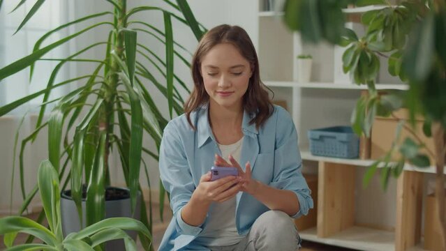 Smiling Young Woman Typing a Message on the Phone among Indoor Plants, Beautiful Young Woman with Beautiful Flowers, a Woman is Holding a Smartphone in her Hands, Stress-Free Life Thanks to Plants