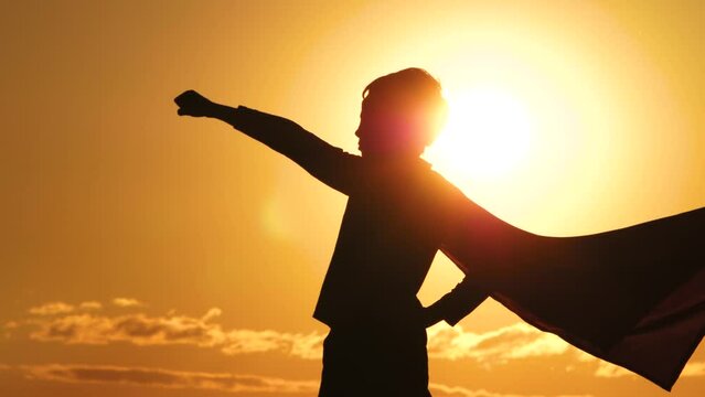 The silhouette of a kid during sunset raises his hand to the sun. Happy superhero boy close-up in a red cape. The concept of a kids dream, strength, success. a red cape fluttering in the wind