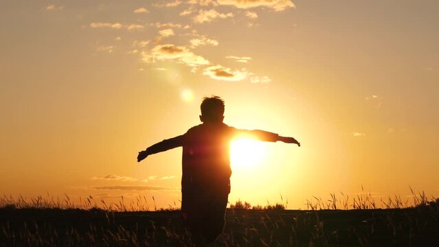 A little kid superhero runs uphill towards the sun during sunset. Silhouette of a happy boy dreaming of becoming a superhero, silhouette of a child from the back. flying hand